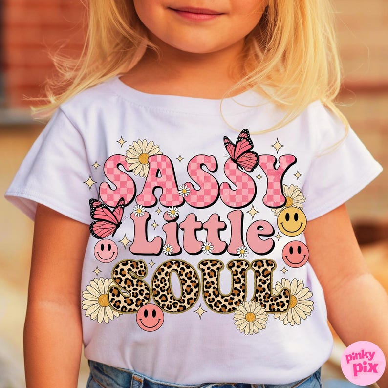 May include: White t-shirt with the text "SASSY Little SOUL" in pink, leopard print, and yellow. The design includes butterflies, daisies, and smiley faces. The shirt is a children's apparel item.