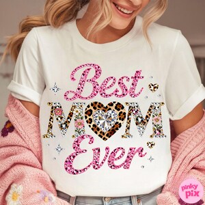 May include: White t-shirt with the text "Best MOM Ever" in pink, floral, and leopard print. The design includes a heart-shaped diamond and decorative stars. The shirt is part of a Mother's Day collection.