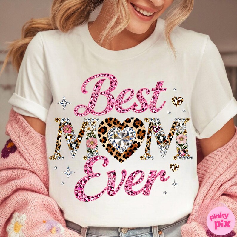 May include: White t-shirt with the text "Best MOM Ever" in pink, floral, and leopard print. The design includes a heart-shaped diamond and decorative stars. The shirt is part of a Mother's Day collection.