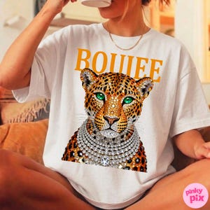 May include: White t-shirt with the word "BOUIEE" in yellow block letters. The shirt features a leopard graphic with green eyes and a diamond necklace. The shirt is a casual, everyday wear item.