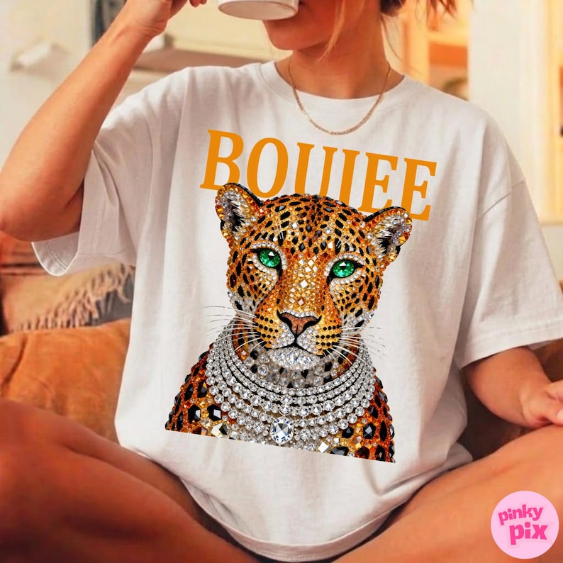 May include: White t-shirt with the word "BOUIEE" in yellow block letters. The shirt features a leopard graphic with green eyes and a diamond necklace. The shirt is a casual, everyday wear item.