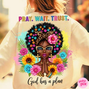 May include: Cream-colored sweatshirt featuring a vibrant graphic of a woman with a floral afro, sunglasses, and praying hands. The design includes the text "PRAY. WAIT. TRUST." and "God has a plan."