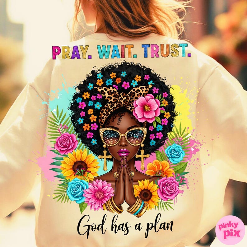 May include: Cream-colored sweatshirt featuring a vibrant graphic of a woman with a floral afro, sunglasses, and praying hands. The design includes the text "PRAY. WAIT. TRUST." and "God has a plan."