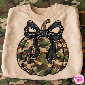 May include: A light tan crewneck sweatshirt featuring a central graphic of a pumpkin with a camouflage pattern in shades of green, brown, and tan. The pumpkin is adorned with a large, black, illustrated bow and outlined in a gold glitter effect.