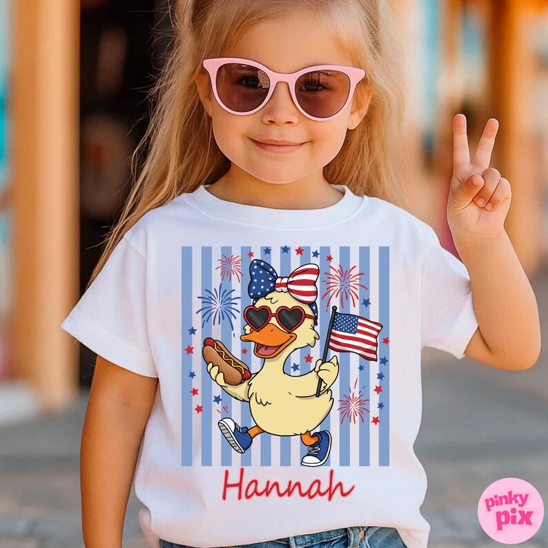 May include: White t-shirt featuring a cartoon duck wearing heart-shaped sunglasses and a patriotic bow, holding a hot dog and an American flag. The design includes fireworks and the name "Hannah" in red.