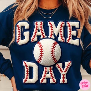 May include: Navy blue sweatshirt with the words "GAME DAY" in white, baseball-themed lettering. A baseball is centered on the sweatshirt. The lettering and baseball have red stitching details. The sweatshirt is a casual style.