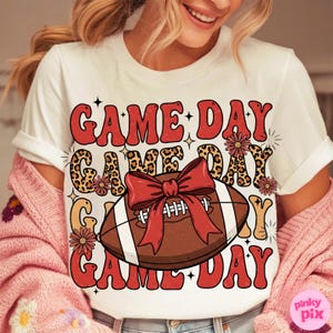 May include: White t-shirt with a retro design featuring the words "GAME DAY" in red, with a football and bow graphic. The design includes leopard print accents and floral details, perfect for game day.