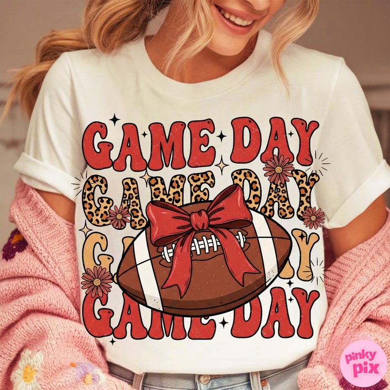 May include: White t-shirt with a retro design featuring the words "GAME DAY" in red, with a football and bow graphic. The design includes leopard print accents and floral details, perfect for game day.