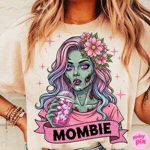 May include: Cream-colored t-shirt featuring a zombie woman with pink and purple hair, wearing a pink shirt and flower crown, drinking from a pink cup. The word "MOMBIE" is on a pink banner. Pink stars are scattered around the graphic.