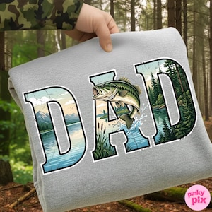 Pu&ograve; includere: Una coperta in pile grigia con la scritta "DAD" a grandi lettere. La "A" presenta un pesce persico che salta e schizzi d'acqua. Le lettere sono riempite con un paesaggio di lago e alberi.
