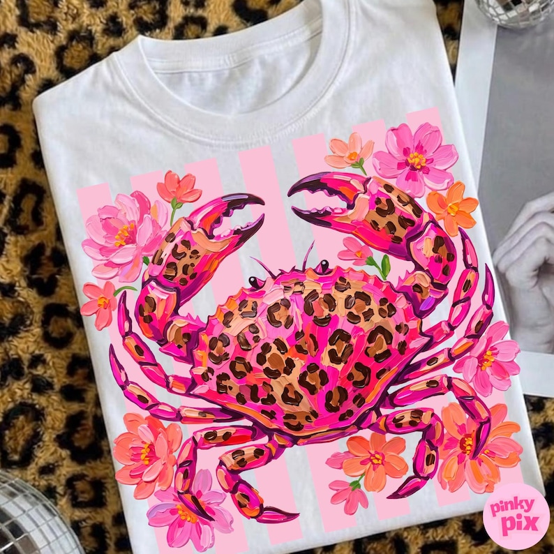 May include: White t-shirt featuring a vibrant crab design. The crab is pink with a leopard print pattern, surrounded by pink and orange flowers. The design is set against a pink striped background.