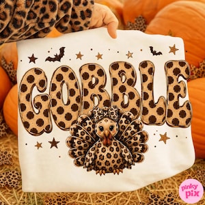 May include: A white t-shirt featuring the word GOBBLE in large, brown, polka-dot letters. Below the text is a cartoon turkey with a leopard-print pattern and a matching bow. The design includes small brown stars and black bat silhouettes.