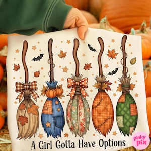 Peut inclure: T-shirt blanc avec cinq balais de sorcière décoratifs avec des feuilles d'automne, des étoiles et des chauves-souris. Chaque balai a un design et un nœud uniques. Le texte "A Girl Gotta Have Options" est imprimé en bas.