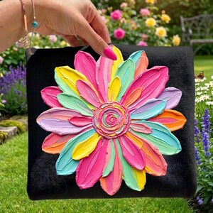 May include: A black square pillow featuring a large, textured, multi-colored flower design. The flower petals are painted in shades of pink, yellow, orange, green, and blue. The pillow is held up in front of a garden.