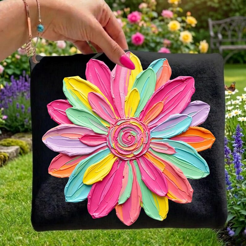 May include: A black square pillow featuring a large, textured, multi-colored flower design. The flower petals are painted in shades of pink, yellow, orange, green, and blue. The pillow is held up in front of a garden.