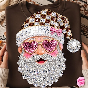 May include: A dark brown t-shirt featuring a graphic of Santa Claus wearing heart-shaped pink sunglasses and a chequered hat. The design is composed of a mosaic of sparkling rhinestones in shades of white, brown, and pink, with a small pink bow.