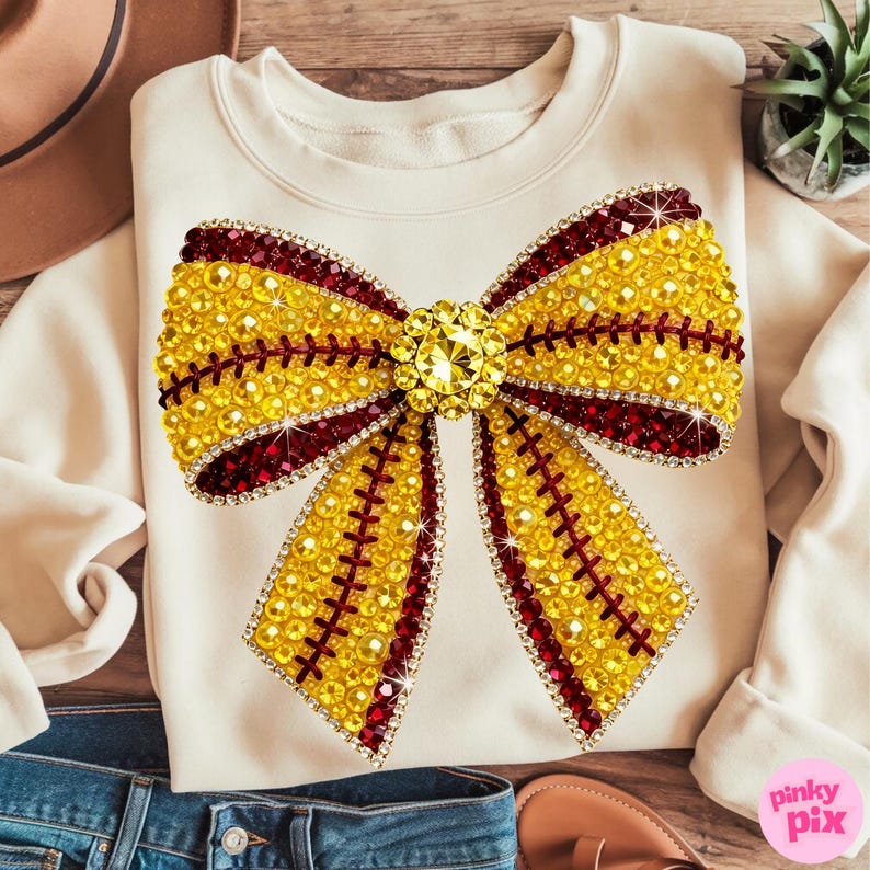 May include: Cream-coloured sweatshirt with a large, embellished bow design. The bow is primarily yellow with red accents and a central jewel. The design is made of rhinestones. The sweatshirt is paired with blue jeans and sandals.
