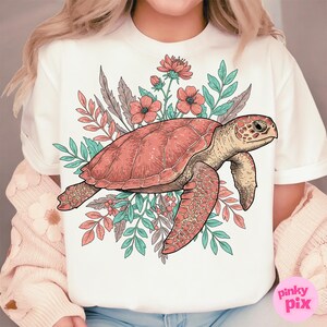 May include: A white t-shirt with a pink and green floral design featuring a sea turtle. The turtle is outlined in black and has a pink shell. The flowers are pink and white with green leaves.