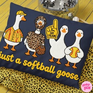 May include: Navy blue pillow with embroidered cartoon geese in baseball and softball gear. The pillow features the text "just a softball goose" in yellow script. A yellow and leopard print fabric is visible.