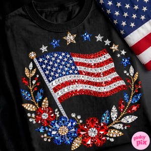 May include: Black t-shirt featuring a sparkling American flag design. The flag is adorned with red, white, and blue crystals, surrounded by a wreath of flowers and stars. A folded American flag is in the background.