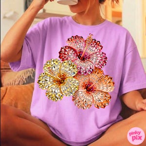 May include: A lilac-colored t-shirt featuring a vibrant floral design. The design showcases three hibiscus flowers in shades of yellow, orange, and red, embellished with sparkling details. The shirt has a relaxed fit.