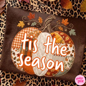 May include: A brown fabric square featuring an embroidered pumpkin design with patchwork patterns in orange, white, and brown. The pumpkin is decorated with autumn leaves and acorns. The text 'tis the season' is printed in white over the pumpkin.