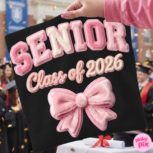 May include: Black graduation cap with the text "SENIOR Class of 2026" in pink and cream-colored lettering. A large pink bow is at the bottom. The cap is held up in front of a graduation ceremony.