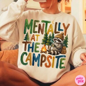 May include: Cream-colored sweatshirt with the text "MENTALLY AT THE CAMPSITE" in colorful block letters. A raccoon wearing a beanie and holding a mug is featured, along with a campfire and trees. The design evokes a camping theme.