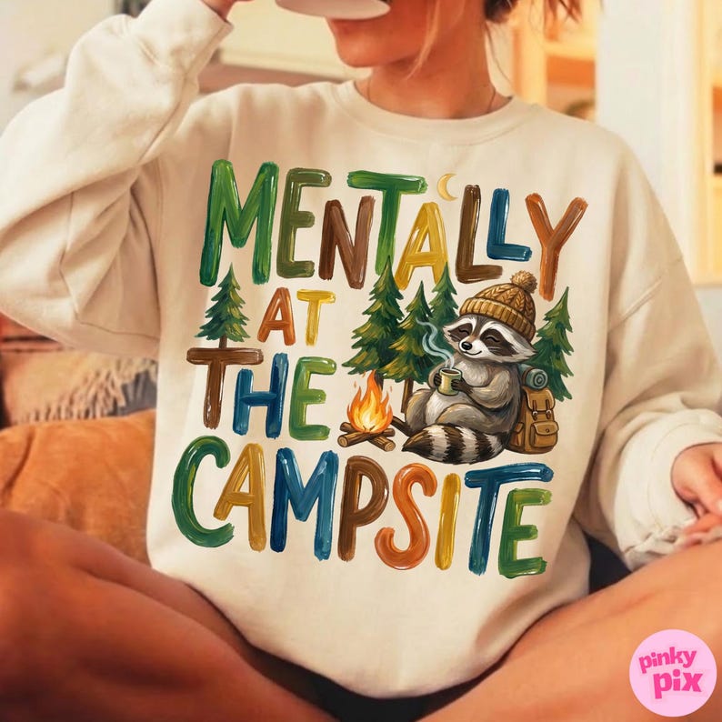 May include: Cream-colored sweatshirt with the text "MENTALLY AT THE CAMPSITE" in colorful block letters. A raccoon wearing a beanie and holding a mug is featured, along with a campfire and trees. The design evokes a camping theme.