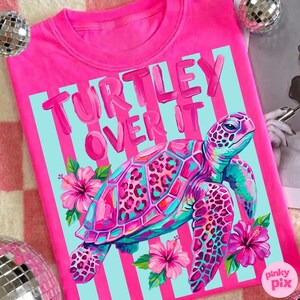 May include: A bright pink t-shirt with the words "TURTLEY OVER IT" in pink lettering. The shirt features a colorful turtle illustration with pink, teal, and purple accents, surrounded by pink hibiscus flowers. The background has light blue stripes.