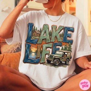 May include: White t-shirt with a colorful graphic that says "LAKE LIFE." The design features a lake scene with a cabin, sunset, and a green jeep. The text is in blue, green, and brown.