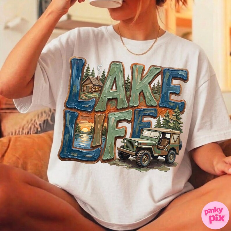 May include: White t-shirt with a colorful graphic that says "LAKE LIFE." The design features a lake scene with a cabin, sunset, and a green jeep. The text is in blue, green, and brown.