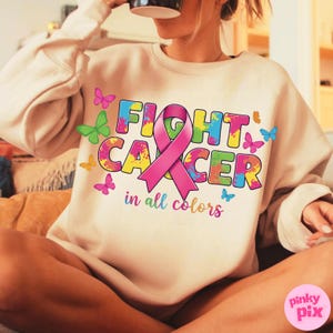 May include: Beige sweatshirt with the words "FIGHT CANCER" in colorful, splattered letters, a pink ribbon, and the phrase "in all colors." Butterflies in various colors are scattered around the design.