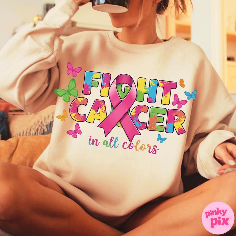 May include: Beige sweatshirt with the words "FIGHT CANCER" in colorful, splattered letters, a pink ribbon, and the phrase "in all colors." Butterflies in various colors are scattered around the design.
