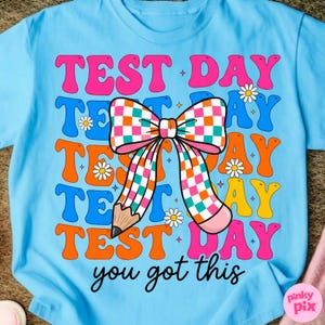 May include: Light blue t-shirt with the words "TEST DAY" in pink, blue, orange, and yellow, with a checkered bow and pencil graphic. The text "you got this" is in black script. Small daisy graphics are scattered around the design.