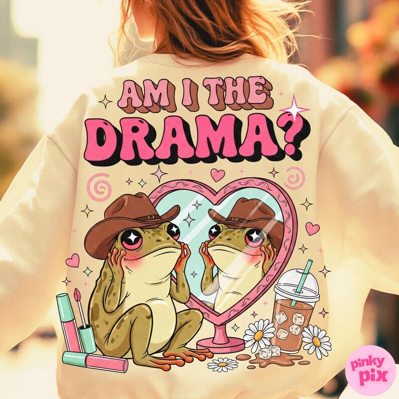 以下が含まれることがあります： ピンク色の「AM I THE DRAMA?」の文字が入ったクリーム色のスウェットシャツ。カウボーイハットをかぶったカエルがハート型の鏡を見ているグラフィック。口紅、デイジー、アイスコーヒーも含まれています。