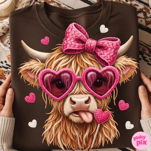May include: Brown sweatshirt featuring a whimsical illustration of a Highland cow wearing pink heart-shaped sunglasses and a pink bow. The cow has its tongue sticking out and is surrounded by pink and white heart accents. The sweatshirt is held by a person.