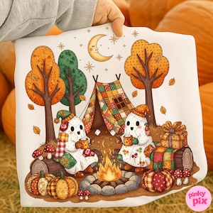 May include: A white sweatshirt featuring a whimsical autumn camping scene. Two ghost figures sit by a campfire, holding mugs. The design includes pumpkins, trees, a tent, and a crescent moon. The overall color palette is warm, with orange, brown, and green hues.