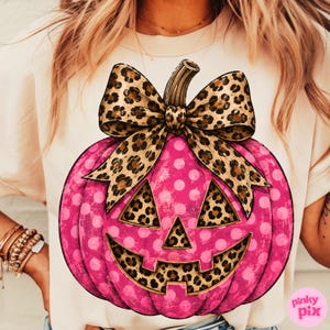 May include: A cream-colored t-shirt featuring a large graphic of a bright pink pumpkin with white polka dots. The pumpkin has a leopard print bow on top and leopard print cutouts for the eyes, nose, and mouth, creating a jack-o'-lantern design.