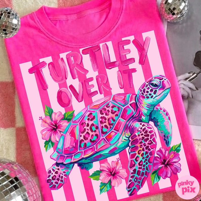 May include: A bright pink t-shirt with the text "TURTLEY OVER IT" in a playful font. The shirt features a colorful illustration of a sea turtle with hibiscus flowers. The turtle has a teal, pink, and purple shell.