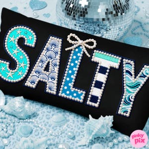 Puede incluir: Cojín rectangular negro con la palabra "SALTY" bordada en turquesa, azul y blanco. Las letras están decoradas con conchas marinas, estrellas de mar, lunares y rayas. La letra "L" tiene un lazo de perlas. Conchas marinas y perlas rodean el cojín.