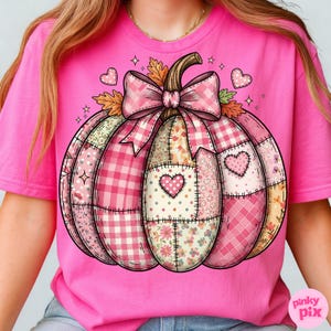 May include: Pink t-shirt featuring a patchwork pumpkin design. The pumpkin is composed of various pink patterns, including gingham, floral, and polka dots, with heart accents. A large pink bow and autumn leaves adorn the top. The shirt has a crew neck.