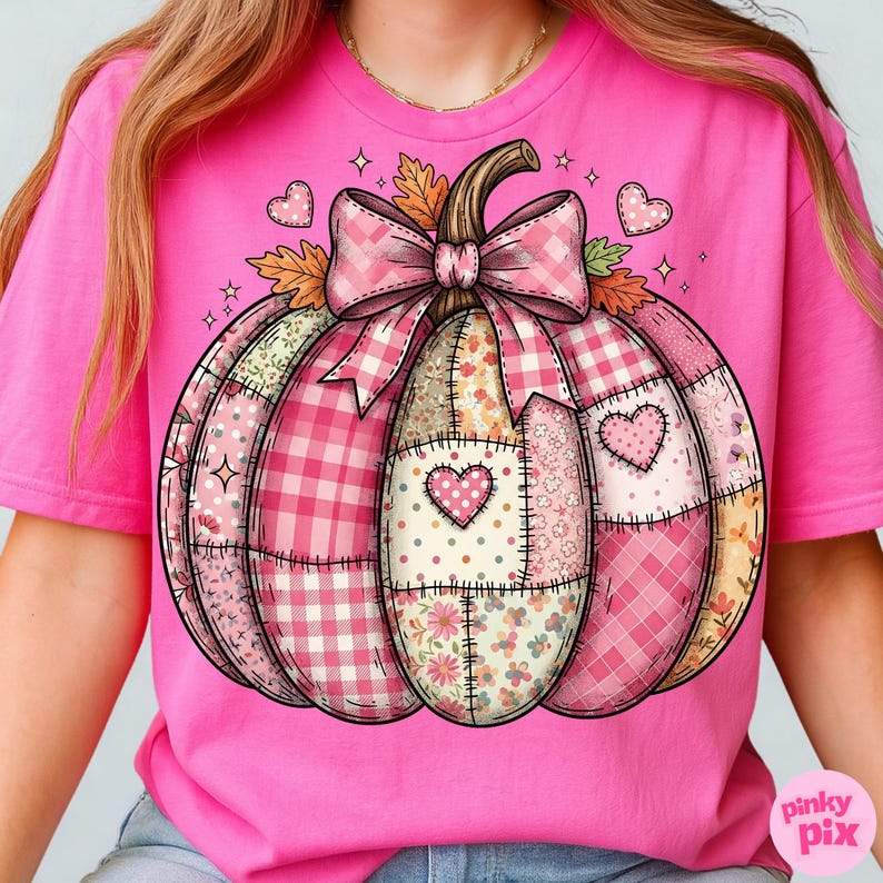 May include: Pink t-shirt featuring a patchwork pumpkin design. The pumpkin is composed of various pink patterns, including gingham, floral, and polka dots, with heart accents. A large pink bow and autumn leaves adorn the top. The shirt has a crew neck.