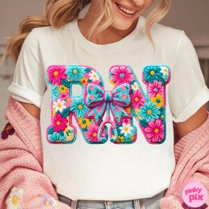 May include: White t-shirt with a colorful floral design spelling "RN". The letters are filled with pink, blue, yellow, and white flowers. A blue bow and stethoscope are in the center. The shirt is a nurse-themed design.