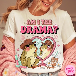 以下が含まれることがあります： ピンクと茶色の「AM I THE DRAMA？」という文字が入った白いTシャツ。カウボーイハットをかぶったカエルがハート型の鏡を見ているグラフィック。口紅、デイジー、飲み物もデザインに含まれています。