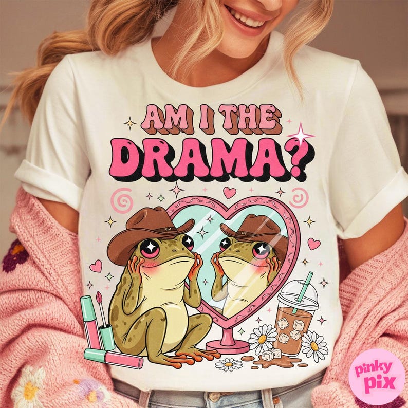 以下が含まれることがあります： ピンクと茶色の「AM I THE DRAMA？」という文字が入った白いTシャツ。カウボーイハットをかぶったカエルがハート型の鏡を見ているグラフィック。口紅、デイジー、飲み物もデザインに含まれています。