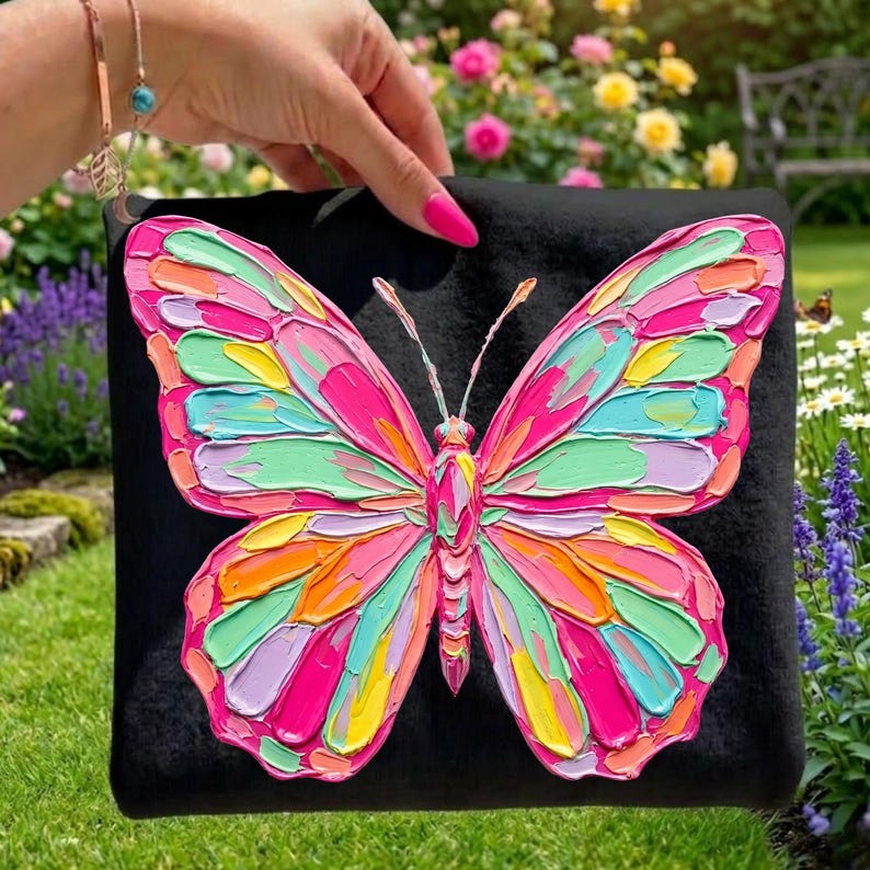 May include: A black clutch featuring a vibrant, textured butterfly design. The butterfly's wings are a mosaic of pink, green, orange, and yellow hues. The artwork is set against a backdrop of a lush garden with colorful flowers.