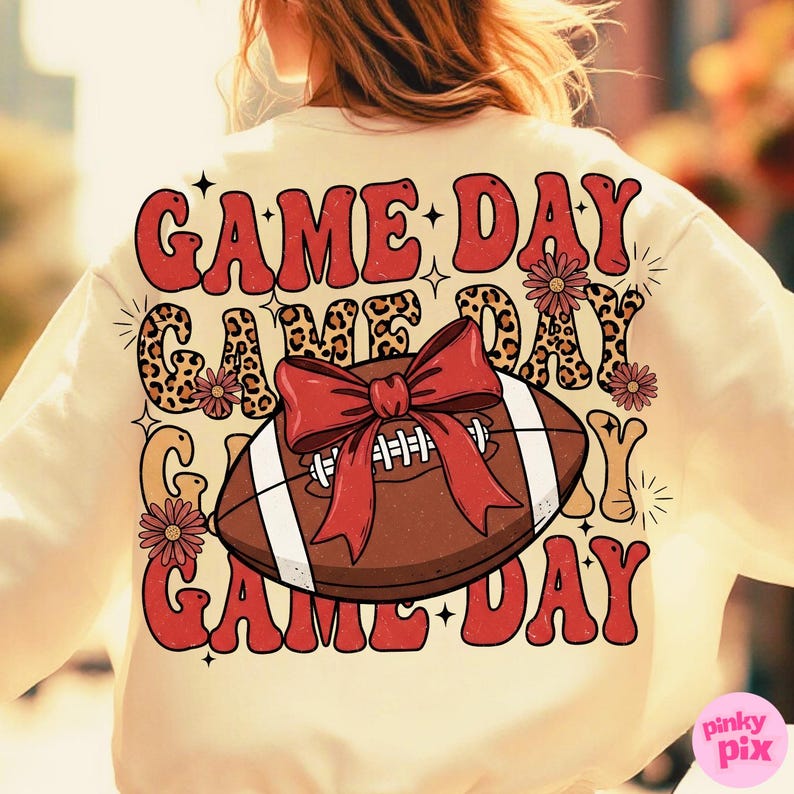 May include: Cream-colored sweatshirt with a football graphic and the text "GAME DAY." The football is brown with white stripes and a red bow. The words "GAME DAY" are in red and leopard print.