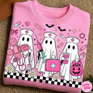 May include: Pink sweatshirt featuring a graphic of three ghost nurses with medical tools and a pumpkin. The design includes a nurse's hat, a stethoscope, a syringe, and a first aid kit. The bottom border has a black and white checkered pattern.
