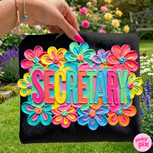 May include: Black decorative pillow with the word "SECRETARY" in colorful 3D lettering, surrounded by vibrant, textured flowers in shades of pink, yellow, blue, and orange. The pillow is held up by a hand.